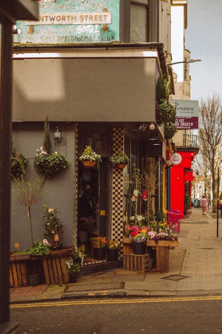 The image depicts the exterior of a florist shop with a welcoming display of fresh flowers arranged outside on wooden crates and hanging baskets. The floral arrangements include bright pink roses, whi