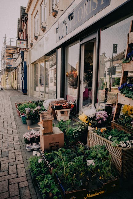 The image depicts the exterior of Fortis Green Flowers, a florist shop with large glass windows and a white facade, showcasing a variety of colorful fresh flowers and plants arranged outside on the pa