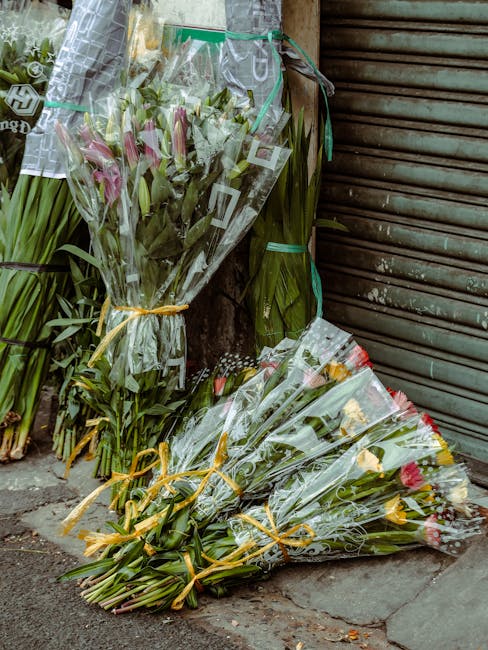 A collection of fresh flower bouquets placed on the sidewalk near a closed, weathered metal shutter. The bouquets include pink lilies and roses, wrapped in clear plastic with yellow ribbons securing t