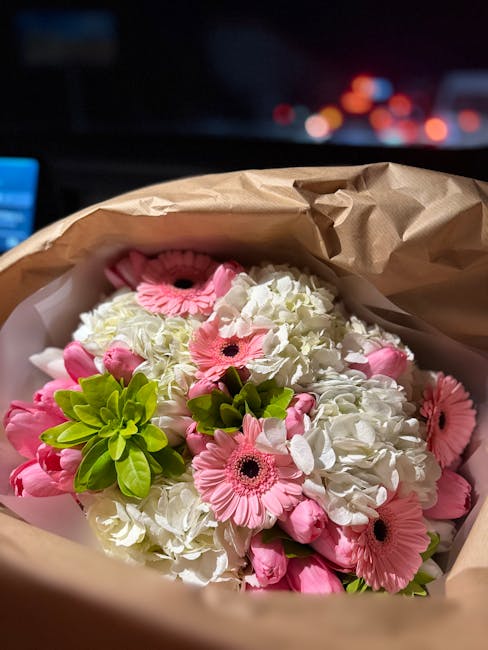 A close-up view of a floral arrangement featuring white hydrangeas and pink gerbera daisies, arranged in a bouquet within brown kraft paper wrapping. The hydrangeas display dense clusters of small, de