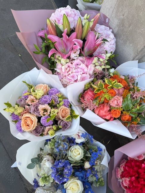A collection of five vibrant floral bouquets displayed on a grey concrete surface, each wrapped in pastel pink or white paper. The top bouquet features large pink lilies, pink hydrangeas, and green fo