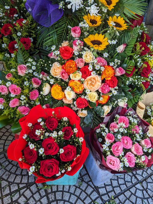 A vibrant display of floral arrangements featuring a variety of fresh flowers with diverse colours and styles. The central bouquet contains a mix of red, pink, yellow, and orange roses, with some whit