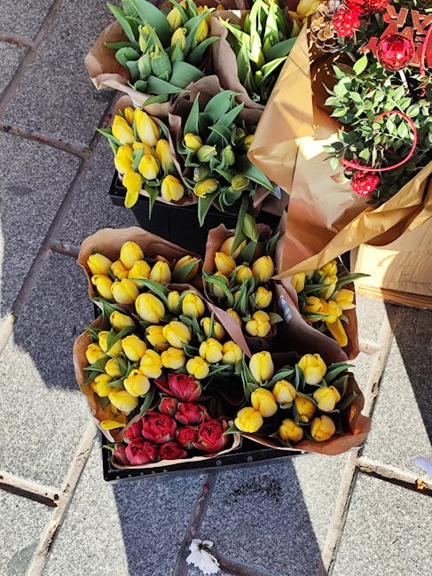 Arrangement of fresh yellow tulips and red roses wrapped in brown craft paper, placed on a stone-tiled surface outdoors. The tulips have vibrant yellow petals with green stems and leaves, while the re