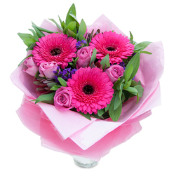 A lively trio of fuchsia Gerbera daisies takes centre stage in this bouquet, each bloom unfurling numerous vivid petals around a deep, shadowed eye that anchors the colour scheme. Interwoven among them are delicate lavender-pink roses in a dusty-rose tone, their petals still snug and velvety as if just opened from the bud, offering a softer, romantic counterpoint to the Gerberas' boldness. Flecks of purple statice drift through the arrangement, providing light, papery texture and a cool contrast that makes the pinks sing. Broad, verdant leaves-likely salal or a hint of eucalyptus-create a verdant backdrop, their rich greens framing the flowers and hinting at the crisp scent of freshly cut foliage. The stems sit inside a clear glass vase whose top rim peeks out beneath an inner wrap of pale pink tissue and an outer layer of translucent fuchsia-pink cellophane; the slightly crinkled cellophane catches the light and gives the gift-ready bouquet a delicate sheen. As a local florist who frequently works around Muswell Hill and Fortis Green, I imagine this composition being delivered for a birthday, anniversary, or a spontaneous celebration, its colours chosen to brighten a kitchen table or a study by the window. The scent is understated-soft rose perfume with a clean green note from the foliage-while the textures range from the satiny rose petals to the crisp, structured Gerbera petals. This arrangement reads as joyful and affectionate, thoughtfully assembled to convey warmth and appreciation on special occasions or quiet moments alike.